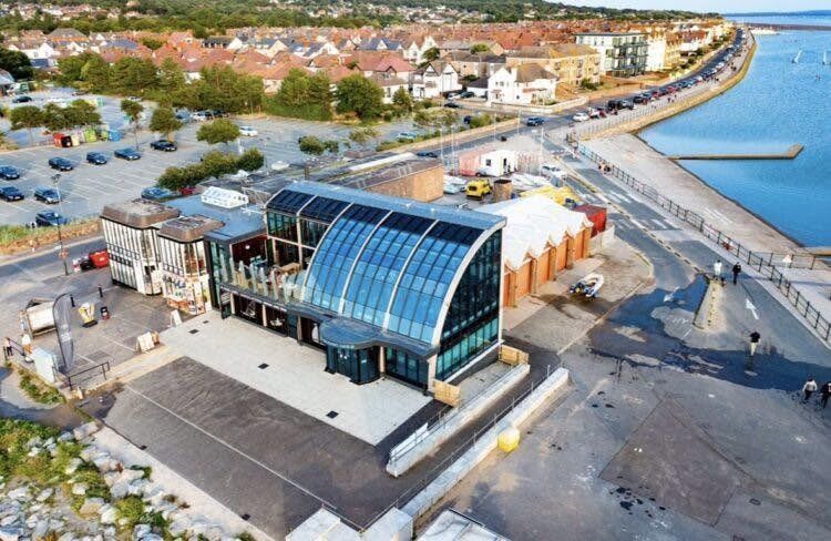 Aerial view of the centre, a curved glass-fronted building beside the car park and promenade at the corner of West Kirby Marine Lake
