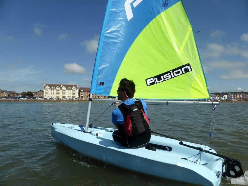 A sailor in a buoyancy aid helming a Fusion dinghy on the marine lake, with the West Kirby seafront behind