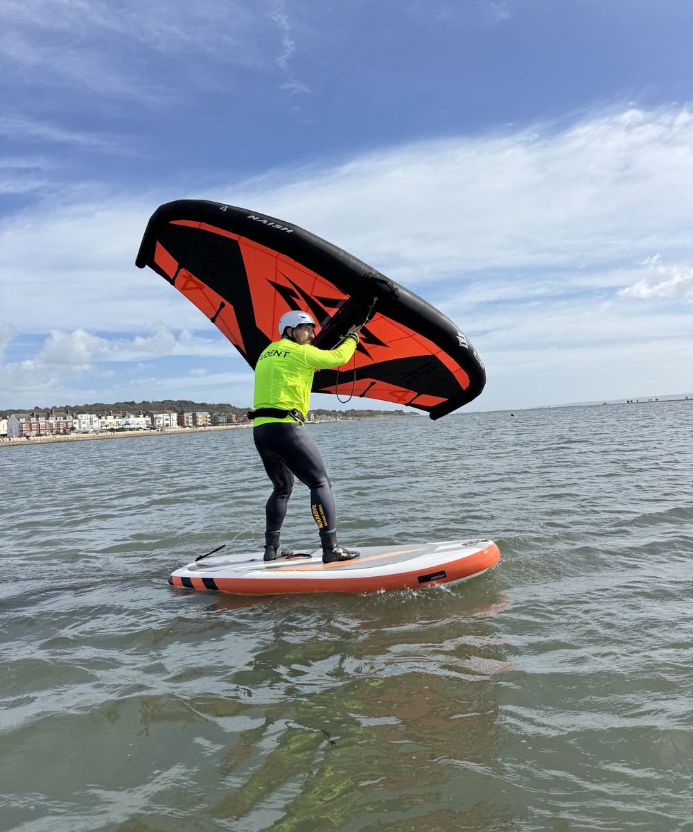 A student on a wing lesson, standing on a board on West Kirby Marine Lake and lifting an orange wing, with the promenade behind