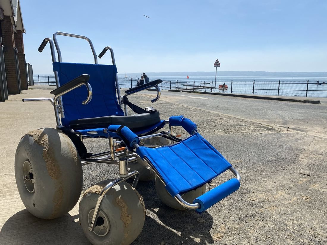 A beach wheelchair with a blue seat and wide balloon tyres outside the centre, with the marine lake and sails behind