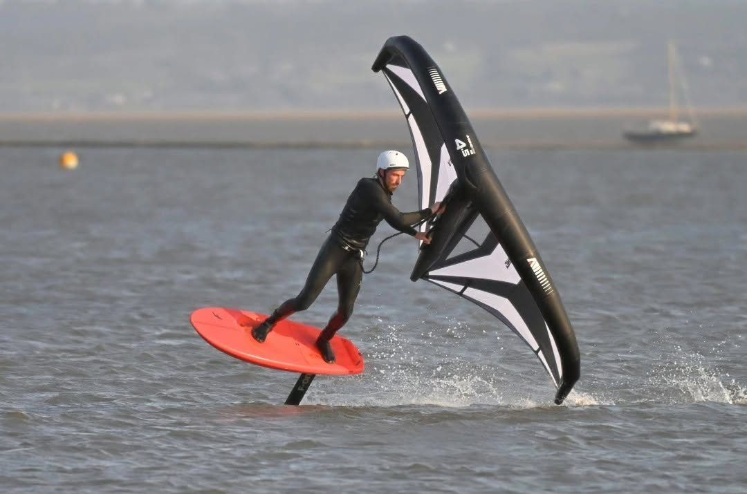 A wing foiler in a white helmet up on the foil on a red board, holding a black and white wing